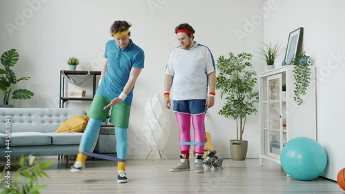 Slow motion of silly guy in bright outfit exercising with stretch band kicking friend having fun indoors in apartment. Sports and humor concept. photo