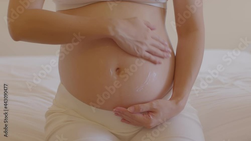 Happy Pregnant Woman sitting on bed holding and stroking apply cream on big belly for beauty moisturizing skin at home,Pregnancy young woman enjoying with future life,Motherhood and Pregnant Concept photo