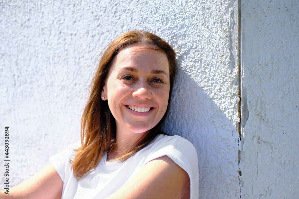 young adult woman smiling and looking at camera. freckled caucasian woman sitting next to an old house wall and enjoying summer