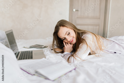 Photo of young joyful woman in pajama with laptop and smiling while sitting on bed in light room