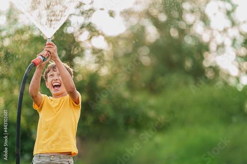 happy boy splashes and plays with garden hose with sprinkler at sunset in backyard, enjoys splashing water in heat. Summer entertainment and outdoor recreation for children. Selective focus on hands.