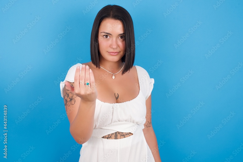 young beautiful tattooed girl wearing white dress standing against blue background, inviting you to come, confident and smiling making a gesture with hand, being positive and friendly. young beautiful tattooed girl wearing white dress standing against blue background, inviting you to come, confident and smiling making a gesture with hand, being positive and friendly.