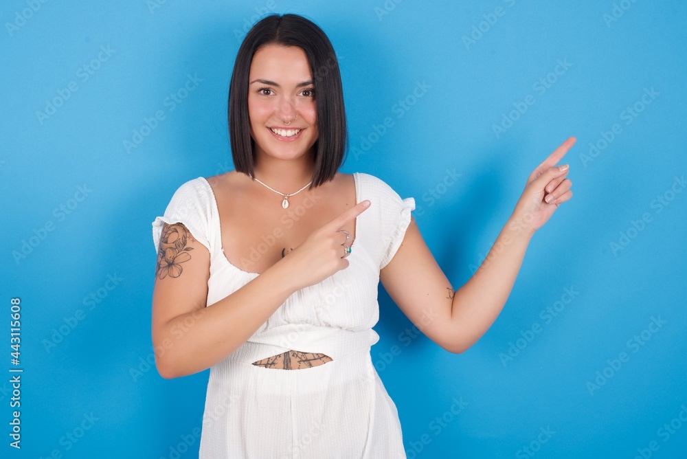 Positive young beautiful tattooed girl wearing white dress standing against blue background with beaming smile pointing with two fingers and looking on empty copy space. Advertisement concept. Positive young beautiful tattooed girl wearing white dress standing against blue background with beaming smile pointing with two fingers and looking on empty copy space. Advertisement concept.