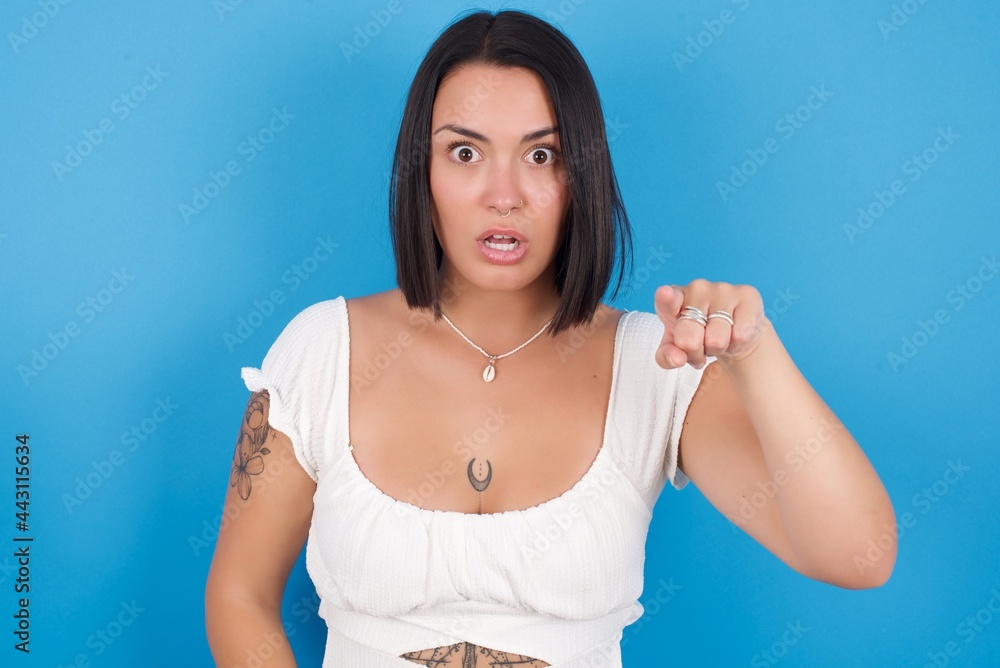 Shocked young beautiful tattooed girl wearing white dress standing against blue background points front with index finger at camera and. Surprise and advertisement concept. Shocked young beautiful tattooed girl wearing white dress standing against blue background points front with index finger at camera and. Surprise and advertisement concept.
