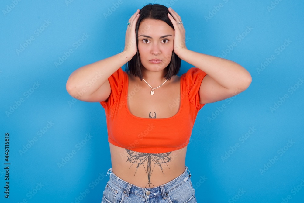 Frustrated young beautiful tattooed girl wearing orange top standing against blue background plugging ears with hands does not wanting to listen hard rock, noise or loud music.