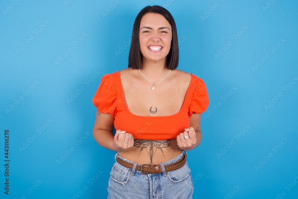 young beautiful tattooed girl wearing orange crop top standing against blue background raising fists up screaming with joy being happy to achieve goals. young beautiful tattooed girl wearing orange crop top standing against blue background raising fists up screaming with joy being happy to achieve goals.