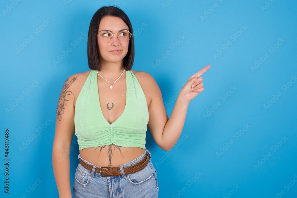 young beautiful tattooed girl wearing green crop top standing against blue background points at copy space and advertises something, advices best price. young beautiful tattooed girl wearing green crop top standing against blue background points at copy space and advertises something, advices best price.