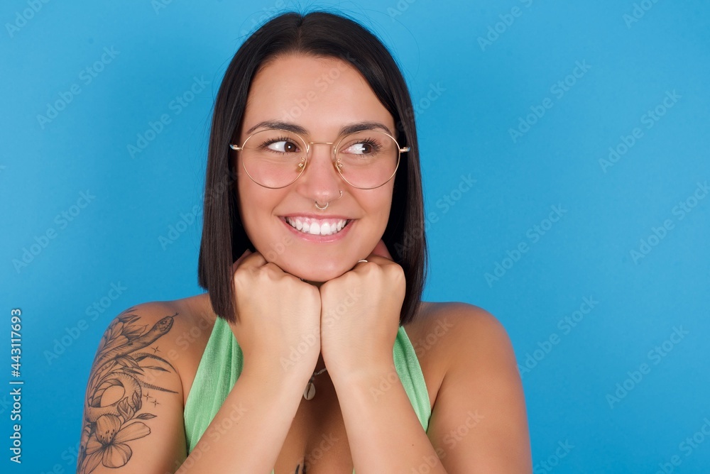 young beautiful tattooed girl wearing green top standing against blue background holds hands under chin, glad to hear heartwarming words from stranger