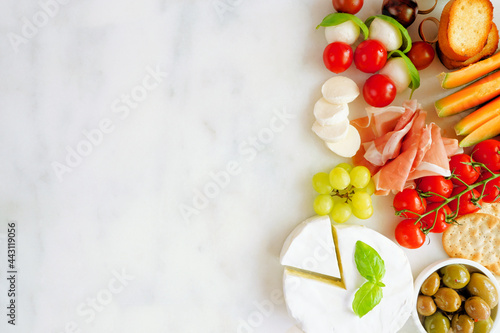 Italian theme charcuterie side border against a white marble background. Selection of cheese, meat and fruit appetizers. Overhead view with copy space.