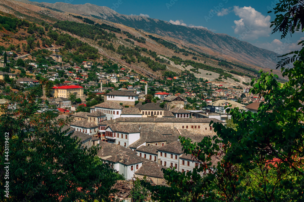  Traditional Albanian  white houses with stone gray roofs and modern buildings on the hill. Summer landscape with lush foliage, blue sky and mountains on the horizon.