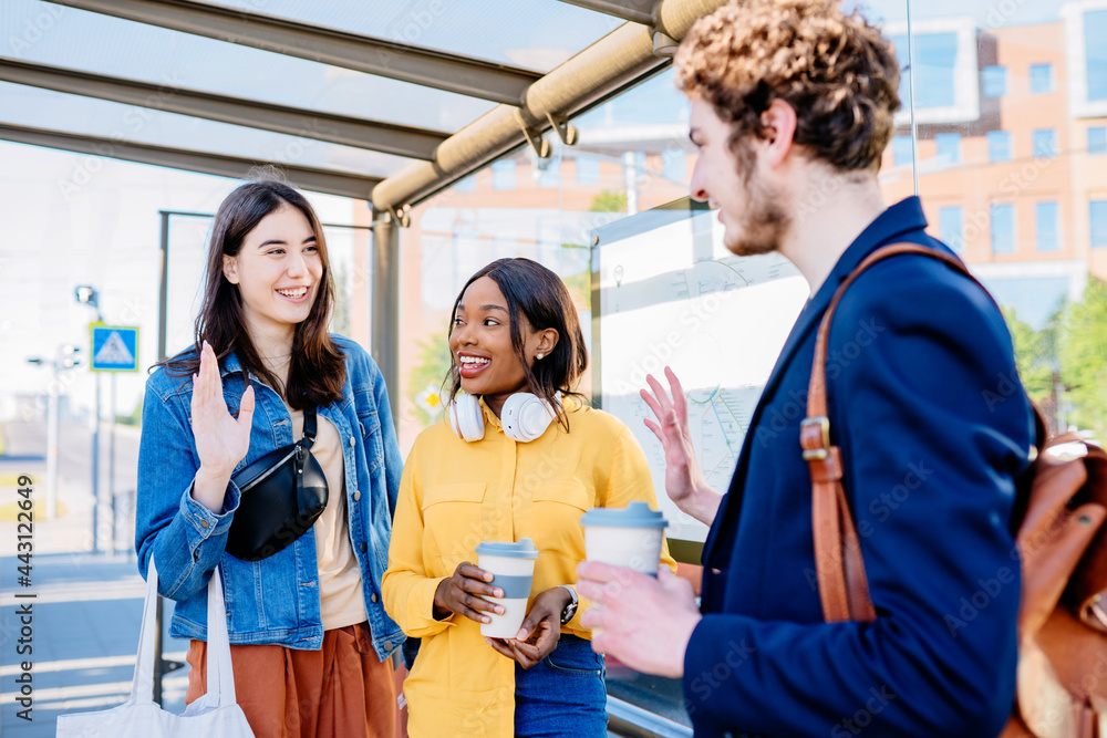 Obraz premium Three multiethnic happy friends meeting in bus stop at street of a big city with an urban background. African and hispanic girl friends student with curly caucasian man commuting to university togethe