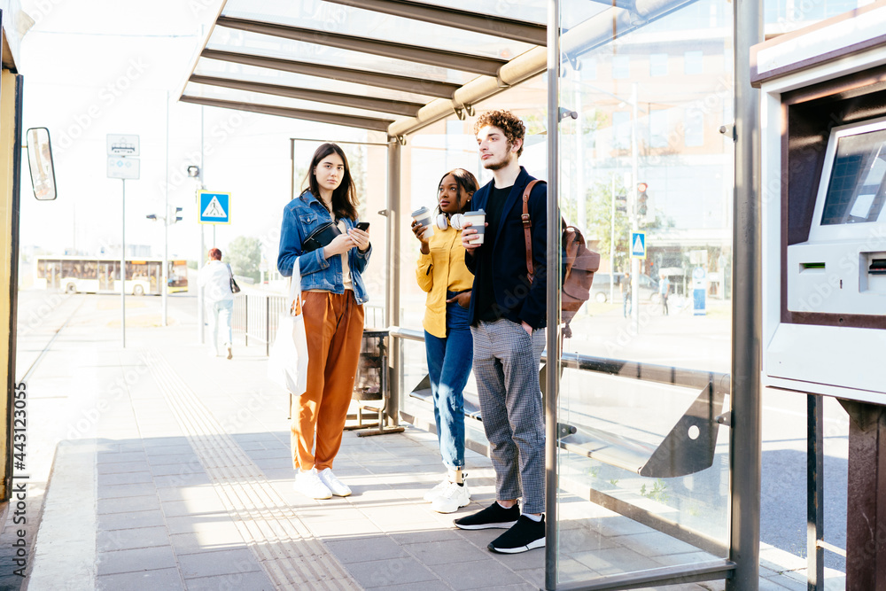 Full height three mixed race friends african women and caucasian man standing at bus stop and waiting for public transport at sunny bright summer day outdoor. African woman with headphones.