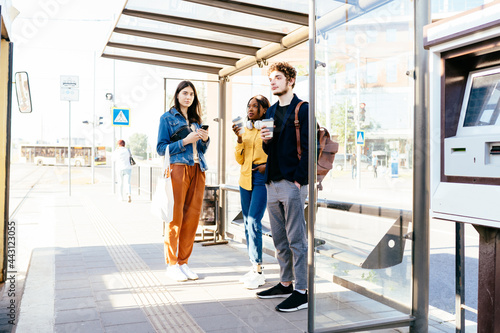 Full height three mixed race friends african women and caucasian man standing at bus stop and waiting for public transport at sunny bright summer day outdoor. African woman with headphones.