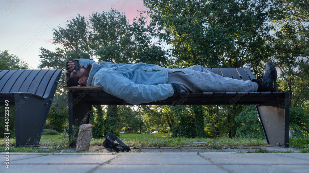 Young tired poor dirty drunk alcoholic homeless man or refugee sleeping on the wooden bench on the urban street in the city, social documentary concept Young tired poor dirty drunk alcoholic homeless man or refugee sleeping on the wooden bench on the urban street in the city, social documentary concept