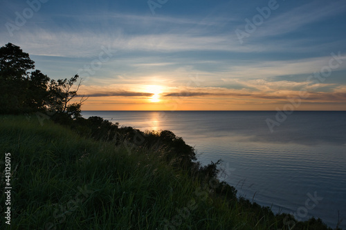 Sunset over the Baltic Sea during the white nights at the shore with a steep slope covered with grass and deciduous forest, under a sky with clouds and haze in the golden tones of the setting sun.