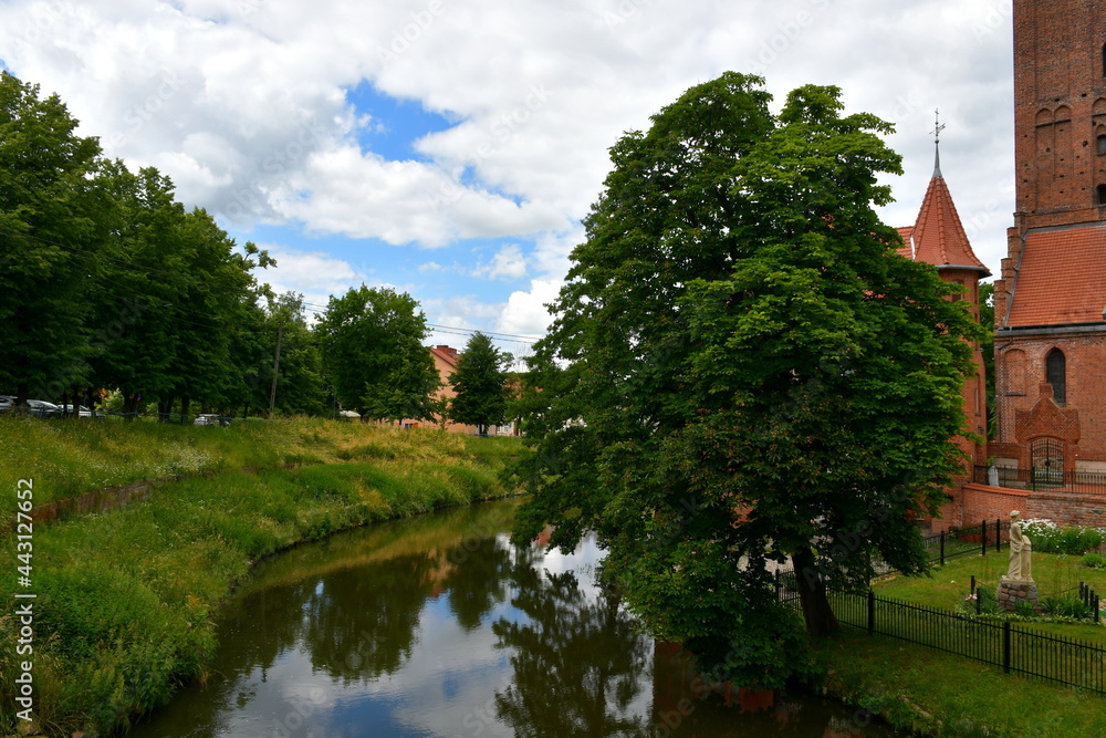 A view of a vast river or lake flowing through a countryside area next to a medieval castle and a church seen next to a public park covered with many deciduous trees spotted on a cloudy summer day