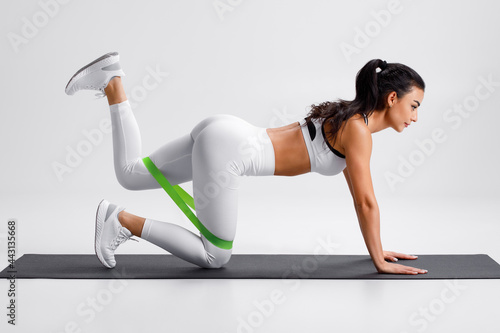 Fitness woman doing kickback exercise for glutes with resistance band on gray background. Athletic girl working out donkey kicks.