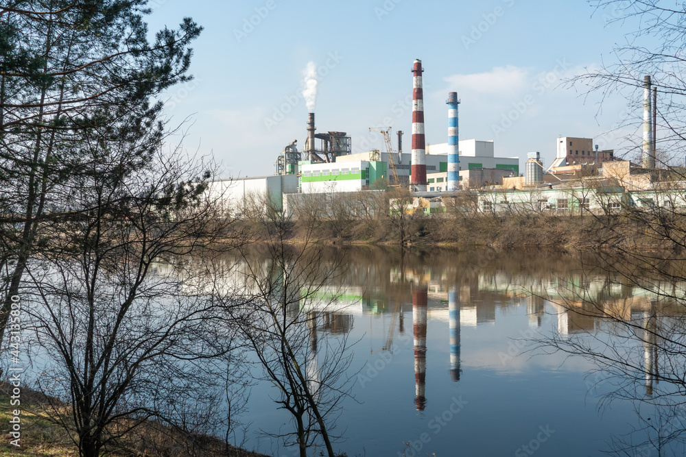 Industrial landscape. A large factory with pipes is reflected in the clear river water against the blue sky. Toxic smoke is coming from the factory's chimney. Environmental pollution