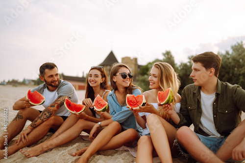 Group of young friends having picnic, eating watermelon, enjoy summer party at the beach. Happy friends resting together sitting near the sea. Beach holiday and summer vacation concept.