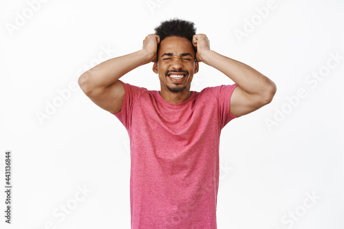 Frustrated african american guy holding hands on head, clench teeth and look distressed, bothered and annoyed by smth, feel complicated, standing against white background
