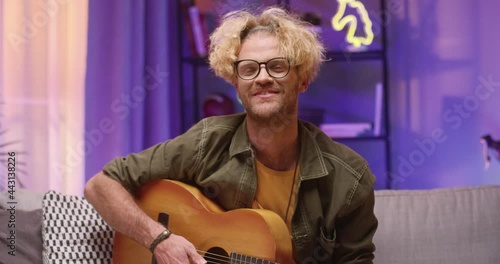 Music podcast. Hipster blonde man holding guitar and playing in microphone in studio while sitting at the sofa alone and creating podcast. Modern mass media and equipment concept photo