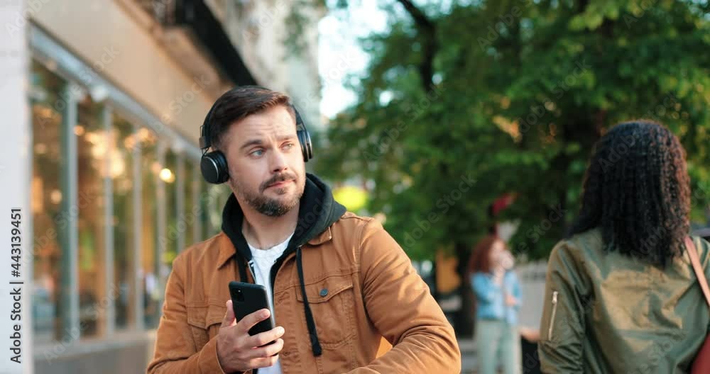 Feeling great. Waist up portrait of the caucasian handsome guy wearing headphones walking through the street and listening music while being in great mood Feeling great. Waist up portrait of the caucasian handsome guy wearing headphones walking through the street and listening music while being in great mood