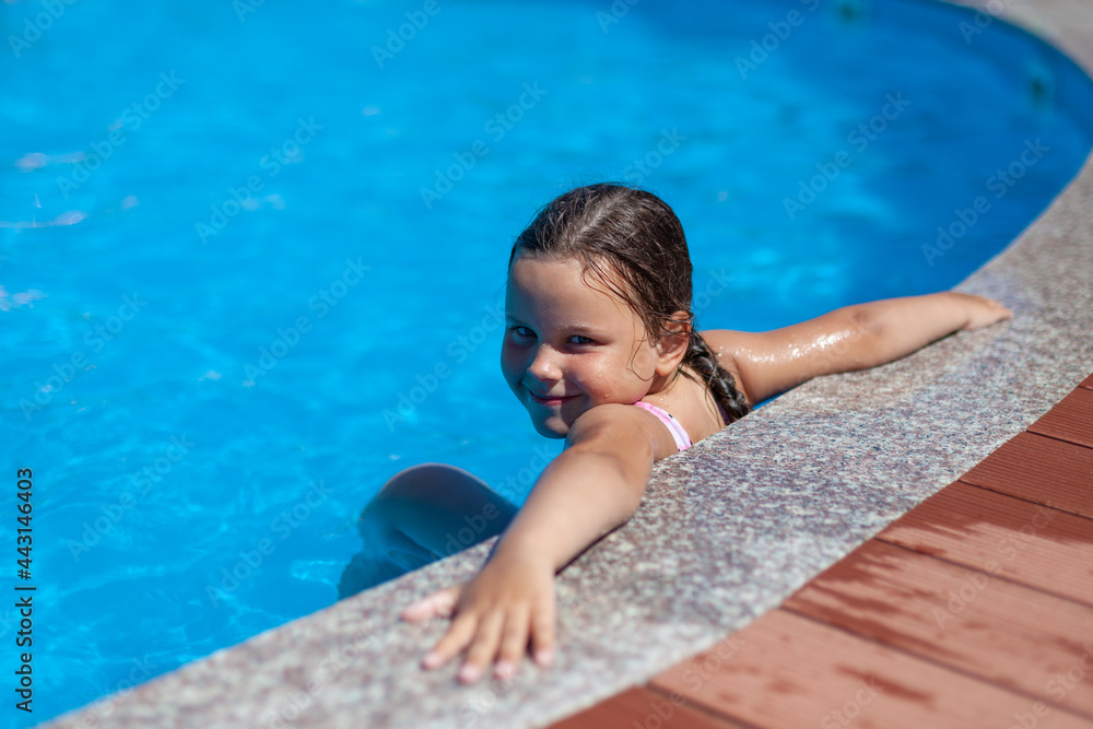 a smiling five-year-old girl put her hands wide on the edge of the pool at the hotel, the concept of a family holiday with children. 