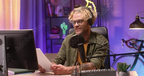 Portrait of creative young man recording podcast and speaking in microphone while holding paper with speech and working at home alone. People and modern lifestyle concept photo