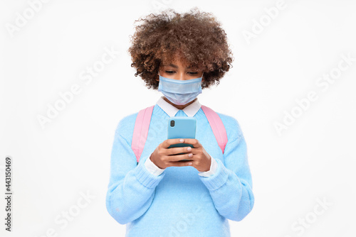 African american teenage girl with afro hairstyle wearing medical mask, holding smartphone with both hands, chatting with friend, isolated on gray background © Damir Khabirov