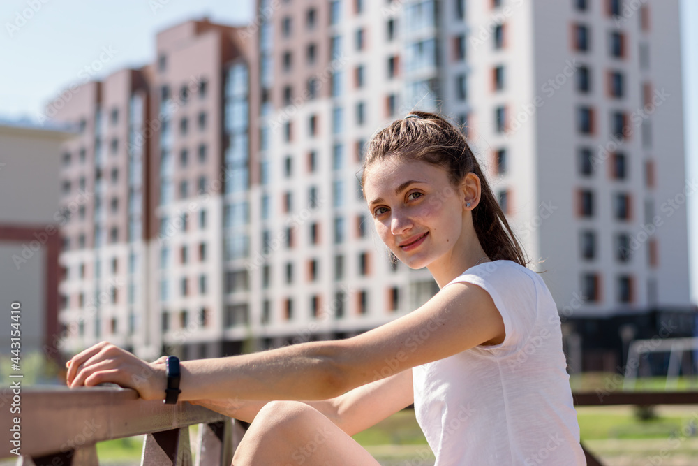 Fototapeta premium A positive young attractive woman in a white T-shirt and with her hair pulled back in a ponytail poses against the background of new high-rise buildings.Summer day.Urban landscape.Copy space.