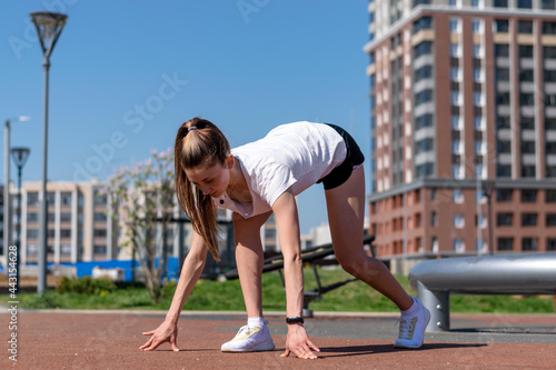 Young athletic woman in a white T-shirt,black shorts and white sneakers .is standing in the low start position before the start of the race on a street sports field.Sports healthy lifestyle.