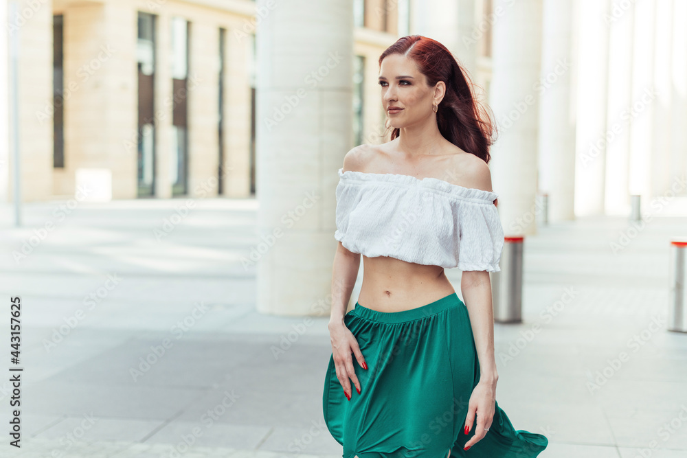 Beautiful red-haired girl model in a blouse and green skirt is walking near a fashionable building Beautiful red-haired girl model in a blouse and green skirt is walking near a fashionable building
