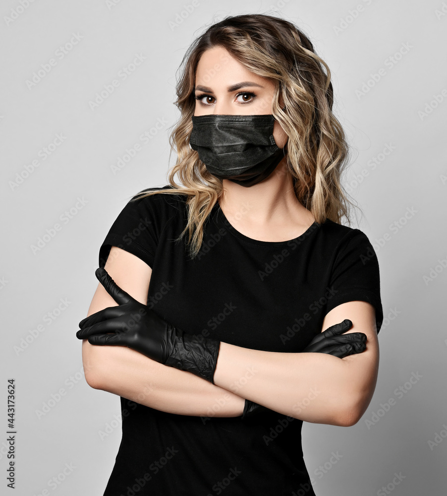 Portrait of blonde curly woman with permanent makeup, eyebrows tattooing in black t-shirt, mask and latex gloves stands holding arms crossed at chest over grey background Portrait of blonde curly woman with permanent makeup, eyebrows tattooing in black t-shirt, mask and latex gloves stands holding arms crossed at chest over grey background