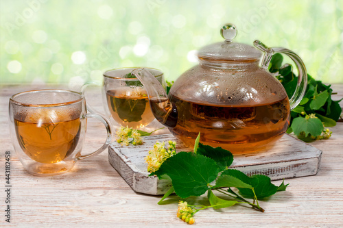 Glass teapot and two cups of tea with a linden tree on a wooden table on a blurred background and linden leaves, flowers. Healing tea.