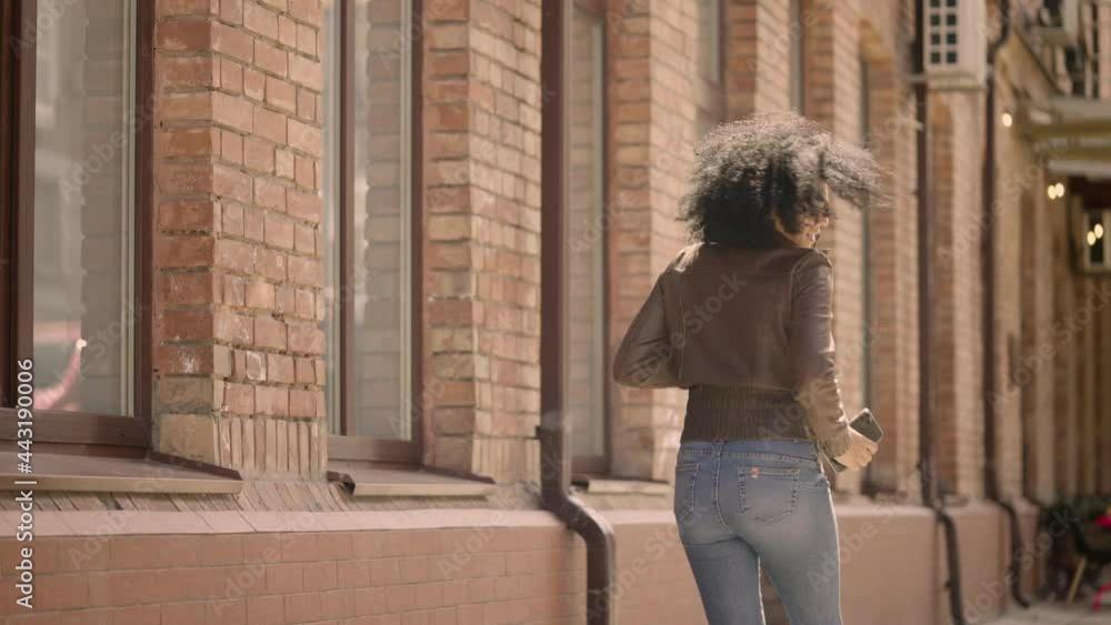 Happy young African American woman walking down street and dancing cheerfully. Brunette with curly hair in brown leather jacket and jeans posing against the backdrop of a brick building. Slow motion. Happy young African American woman walking down street and dancing cheerfully. Brunette with curly hair in brown leather jacket and jeans posing against the backdrop of a brick building. Slow motion.