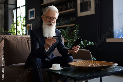 Successful businessman sitting on sofa in office and pays by credit card with his mobile phone.