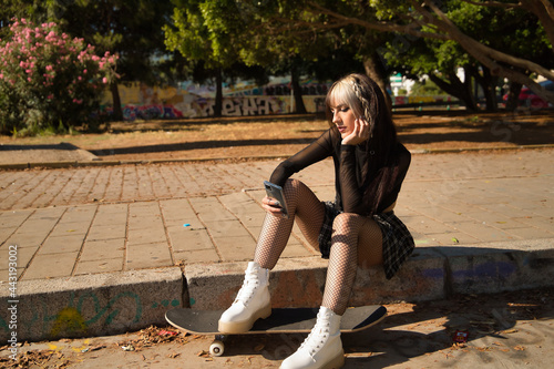 Cute young girl with heterochromia and punk style in black blouse and plaid skirt sitting on a curb with her feet on a skateboard checking social media on her phone.