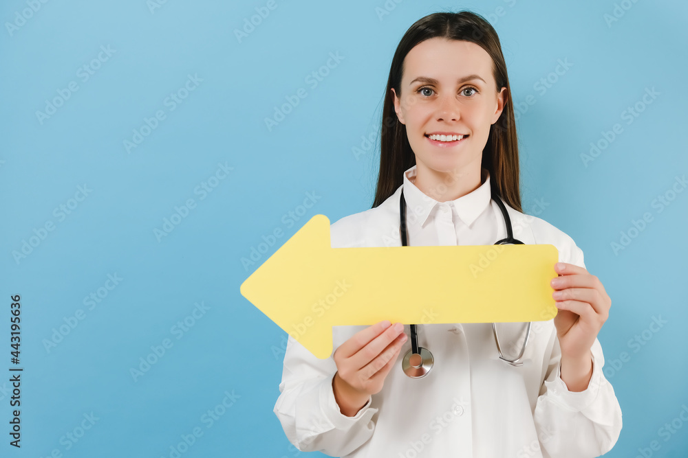 Smiling medical female doctor in white medical uniform pointing direction with yellow arrow aside, isolated over blue studio background wall. Healthcare personnel medicine concept. Mock up copy space Smiling medical female doctor in white medical uniform pointing direction with yellow arrow aside, isolated over blue studio background wall. Healthcare personnel medicine concept. Mock up copy space