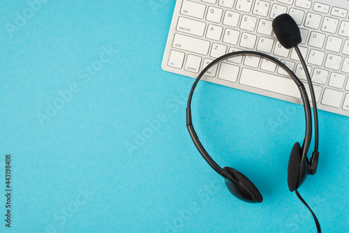 Top view photo of black headphones with microphone and white keyboard on isolated light blue background with copyspace