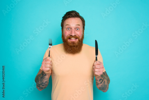 Quadro em tela Happy man with tattoos is ready to eat with cutlery in hand