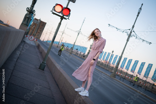 Portrait of a beautiful girl of European appearance in a retro dress stands on a drawbridge on the road with red traffic light.