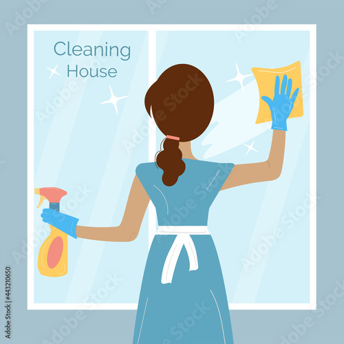 Woman in a blue uniform is washing the window with duster, holding a spray in her hands
