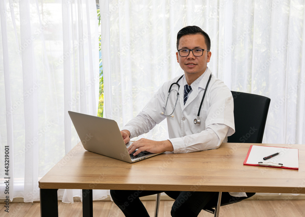 Asian young male happiness doctor typing laptop sitting at desk in hospital room Asian young male happiness doctor typing laptop sitting at desk in hospital room