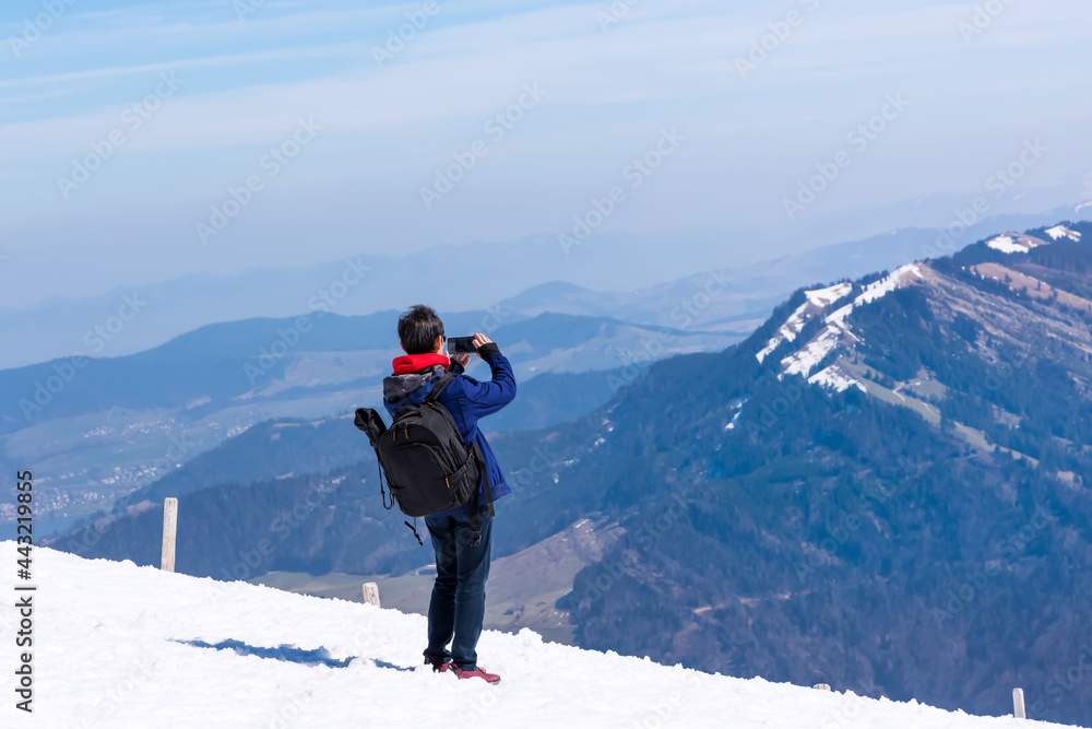 Back view of young tourist blogger travel with snow peak view and  taking photo by smartphone with snow mountain at Switzerland with blue sky background