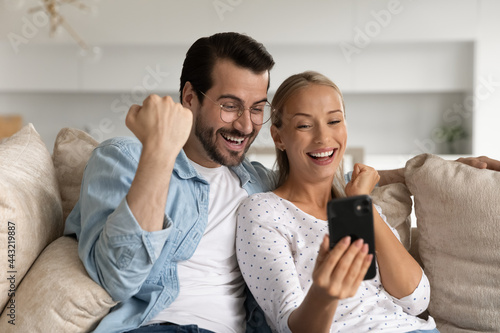 Excited young couple looking at phone screen together, celebrating success close up, sitting on couch, overjoyed woman and man in glasses rejoicing online lottery win, reading unexpected good news