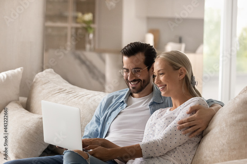 Happy wife and husband in glasses hugging, relaxing on couch with laptop, smiling young couple looking at computer screen, shopping or chatting in social network together, having fun online