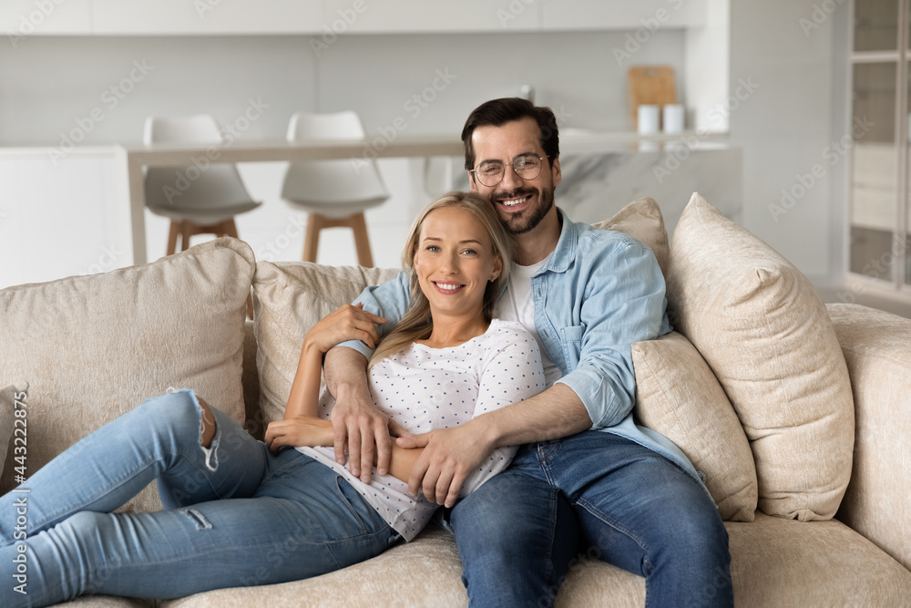 Portrait of smiling young couple hugging sitting on comfortable couch at home together, happy beautiful wife and husband in glasses looking at camera, posing for family photo in modern living room