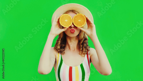Summer portrait of cheerful young woman covering her eyes with slices of orange fruits and looking for something wearing a straw hat on green background