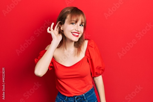 Redhead young woman wearing casual red t shirt smiling with hand over ear listening an hearing to rumor or gossip. deafness concept.