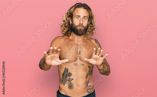 Handsome man with beard and long hair standing shirtless showing tattoos moving away hands palms showing refusal and denial with afraid and disgusting expression. stop and forbidden.